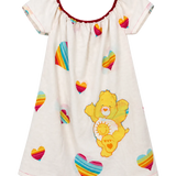 A short-sleeve baby dress or nightgown in off-white, featuring a colorful rainbow-striped heart pattern and a large yellow Care Bear character print on the front. The dress has a red-trimmed scoop neckline and is shown against a plain white background.