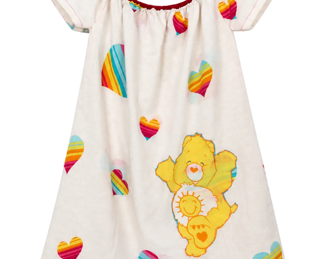 A short-sleeve baby dress or nightgown in off-white, featuring a colorful rainbow-striped heart pattern and a large yellow Care Bear character print on the front. The dress has a red-trimmed scoop neckline and is shown against a plain white background.