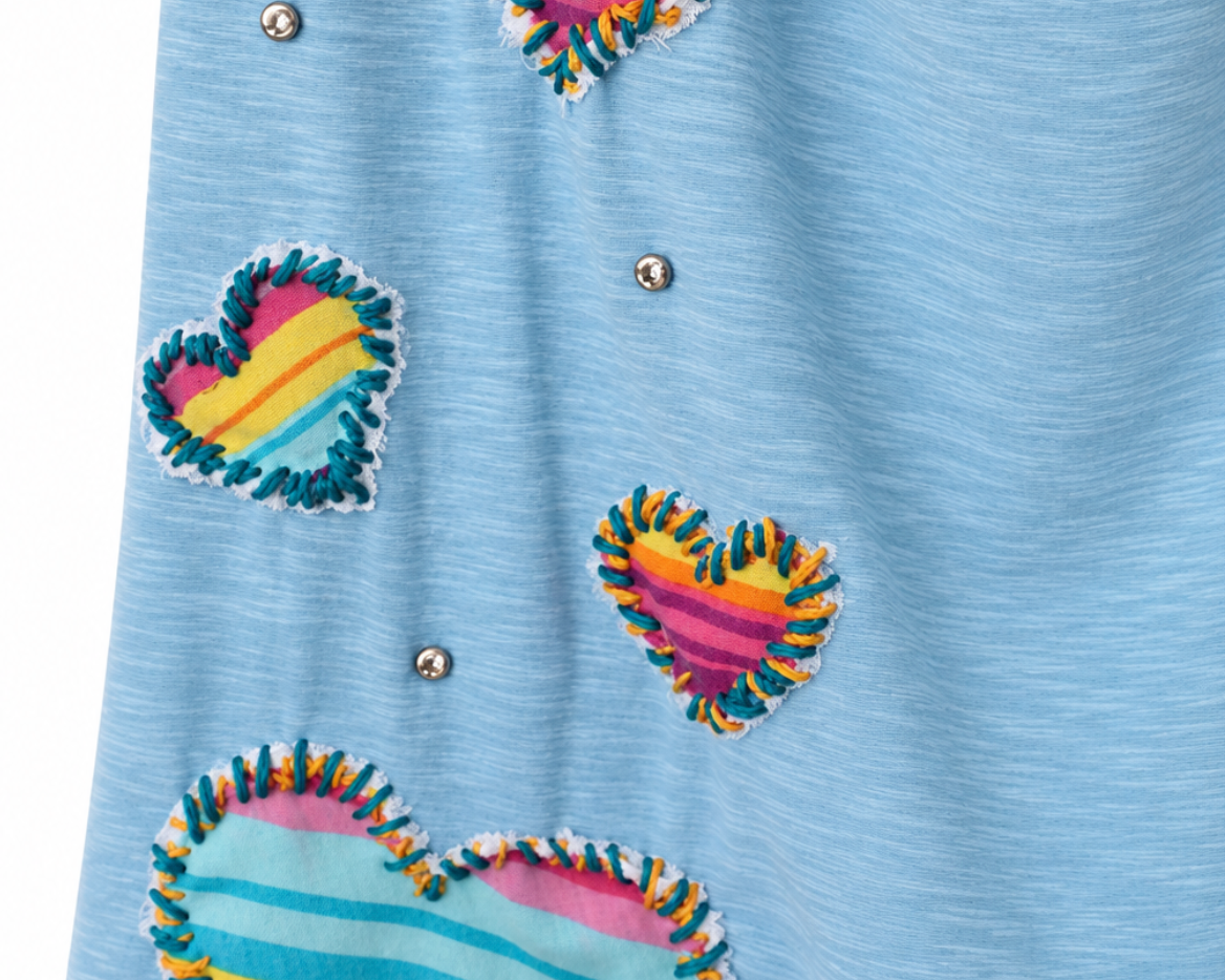 A close-up of a light blue heathered fabric garment featuring three colorful striped fabric hearts with frayed edges and small silver rhinestone embellishments.
