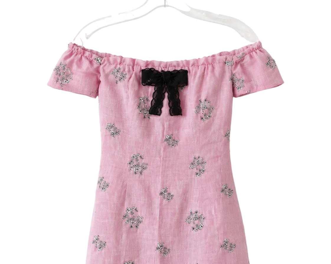 A pink off-the-shoulder dress with a black bow at the neckline and small floral embroidery, hanging on a clear plastic hanger against a white background.