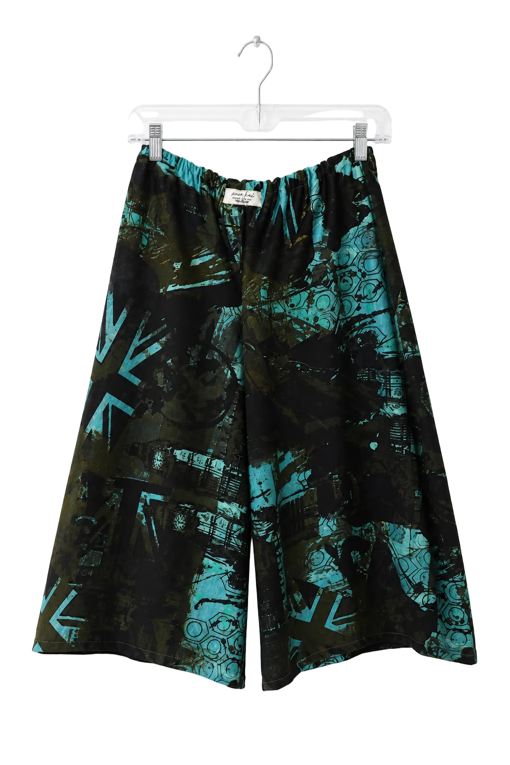 A pair of wide-leg shorts with an abstract teal and black graffiti-style print, hanging from a clear plastic hanger against a plain white wall.
