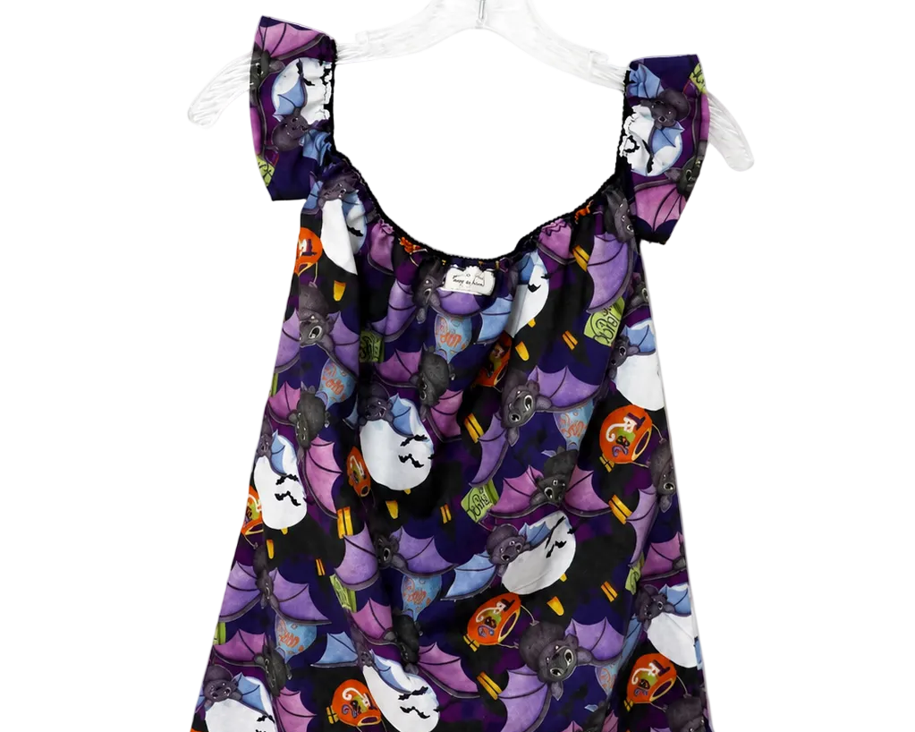 A sleeveless babydoll dress with a dark purple and black background, featuring a playful print of cartoon cats in white, purple, orange, and yellow. The dress has ruffled shoulder straps and is displayed on a clear plastic hanger against a plain white background.