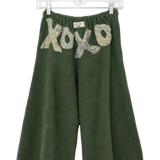 A pair of dark green wide-leg cropped pants hanging on a clear plastic hanger against a white background. The pants feature a textured, snakeskin-patterned 'xo xo' graphic across the upper front.