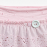 Close-up of a light pink eyelet skirt with a ruffled waistband, featuring three decorative buttons in pink and white.