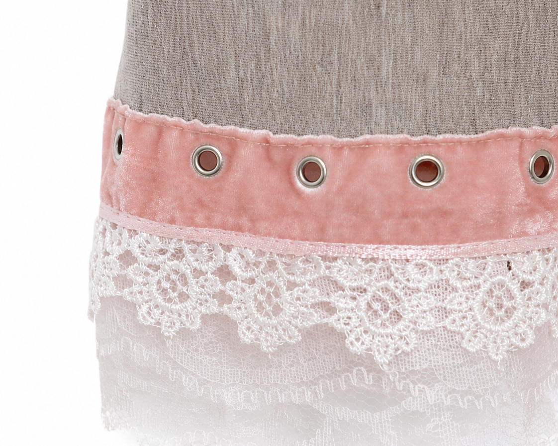 Close-up of a skirt or dress hem featuring a dusty pink velvet band with silver eyelets, layered over a white floral lace trim, on a light gray textured fabric.