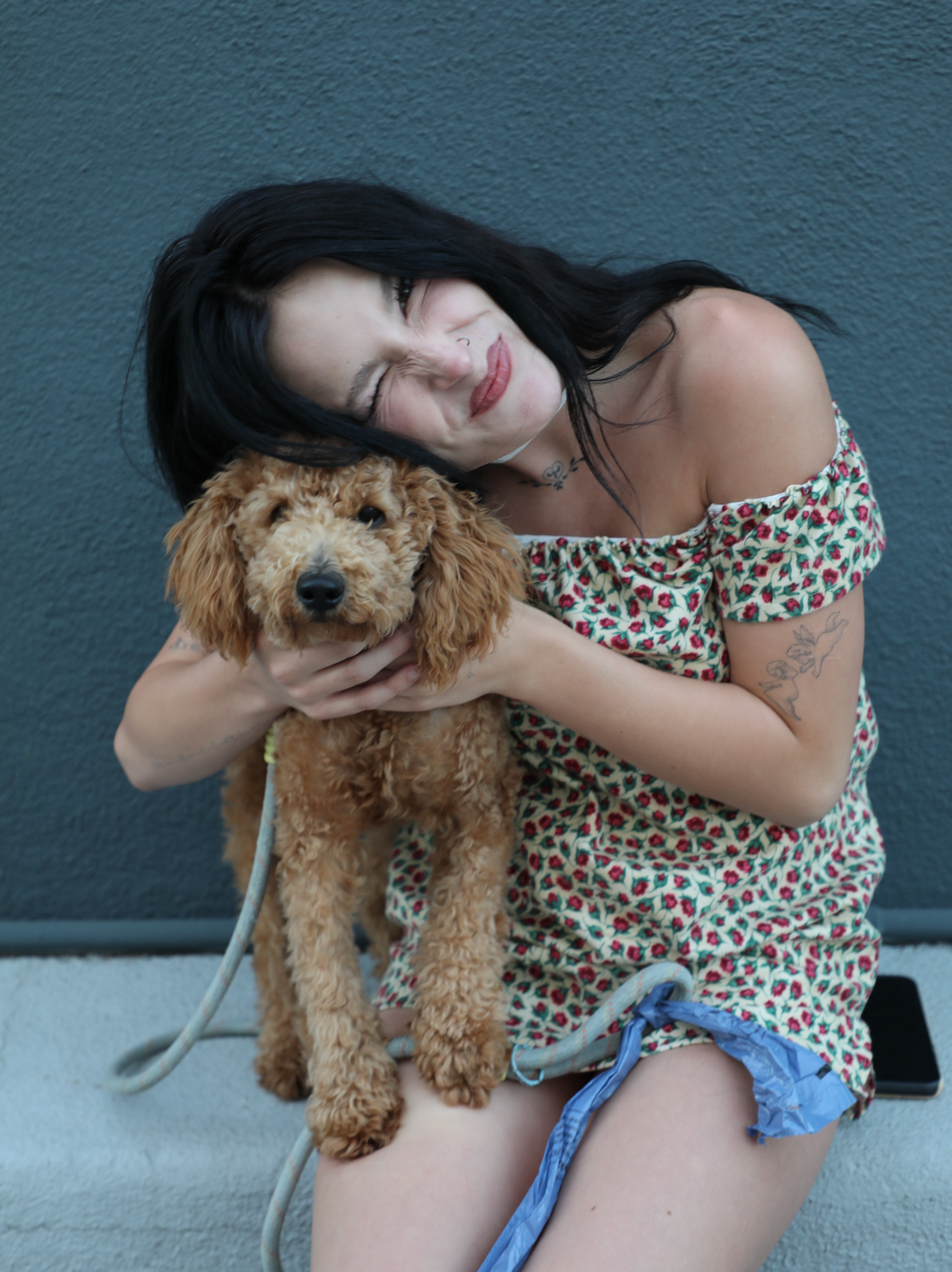A Kaiwith dark hair wearing a cherry-patterned off-the-shoulder top and denim shorts sits on a concrete surface, holding a small brown poodle dog against a dark blue-gray wall.
