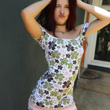 A Kai wearing the Margo Dress. white scoop-neck mini dress with a green, brown, and pink clover pattern, lace trim, and a ruffled hem, posing outdoors against a dark gray wall.