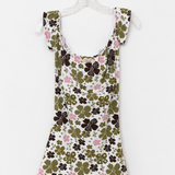 A sleeveless white babydoll dress with a green and brown clover pattern, pink floral accents, and a pink lace hem, hanging on a white plastic hanger against a white background.