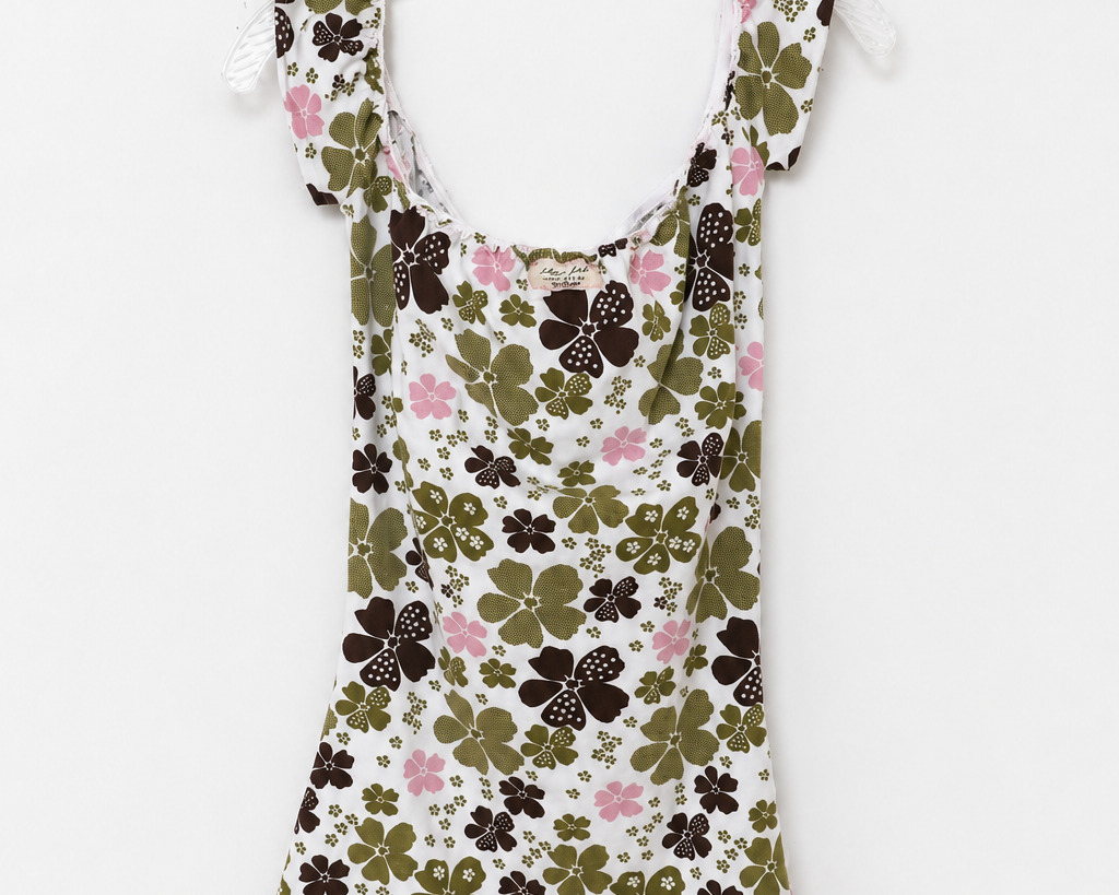 A sleeveless white babydoll dress with a green and brown clover pattern, pink floral accents, and a pink lace hem, hanging on a white plastic hanger against a white background.