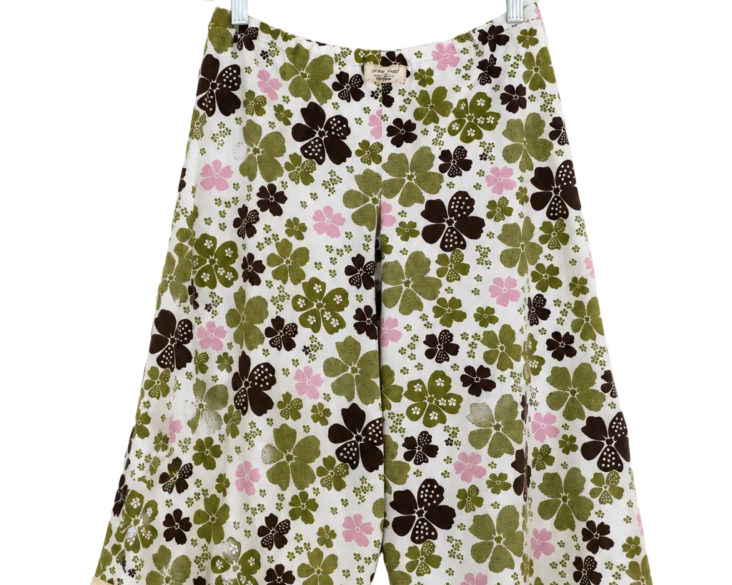 A pair of women's wide-leg shorts with a retro floral pattern featuring olive green, dark brown, and pink flowers on a cream background, hanging on a clear plastic hanger against a white wall.