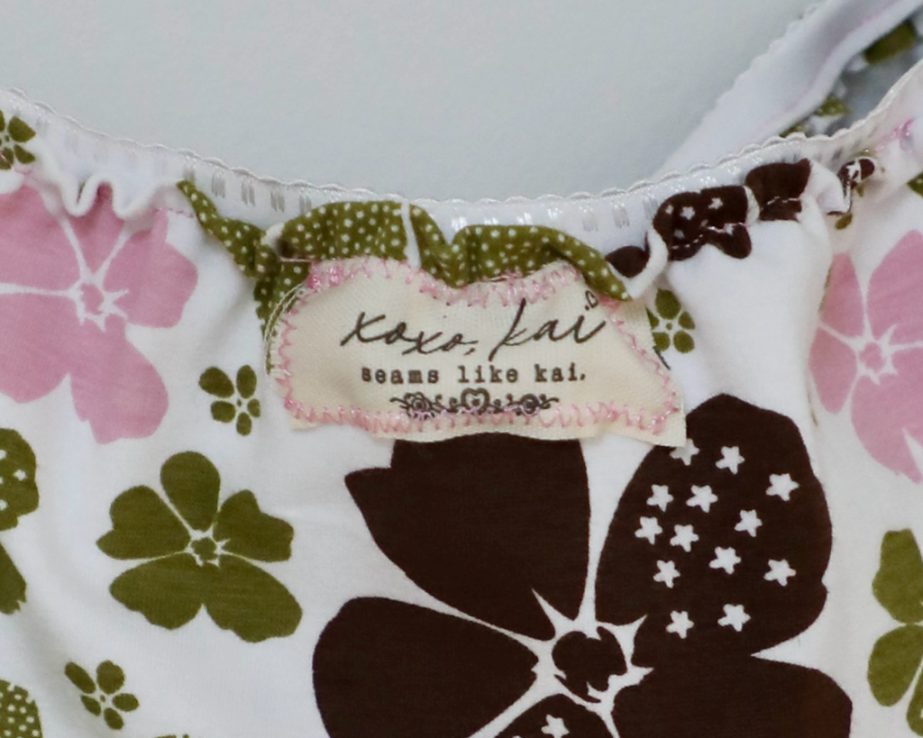 Close-up of a floral-patterned garment, likely a bikini top, featuring a white base with large dark brown and olive green flowers, pink floral accents, and a visible 'Koko Kai' brand label.