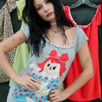 A Kai modeling a gray short-sleeve t-shirt dress featuring a large Raggedy Ann doll graphic on the front, posed in front of a clothing rack with other garments.