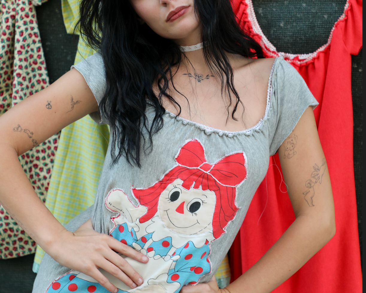 A Kai modeling a gray short-sleeve t-shirt dress featuring a large Raggedy Ann doll graphic on the front, posed in front of a clothing rack with other garments.