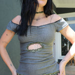 Kai modeling a gray off-the-shoulder top with a central cutout framed by green chain detailing, paired with matching gray shorts featuring a circular patch, photographed outdoors against a building wall.