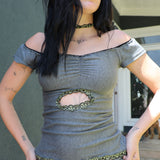Kai modeling a gray off-the-shoulder top with a central cutout framed by green chain detailing, paired with matching gray shorts featuring a circular patch, photographed outdoors against a building wall.