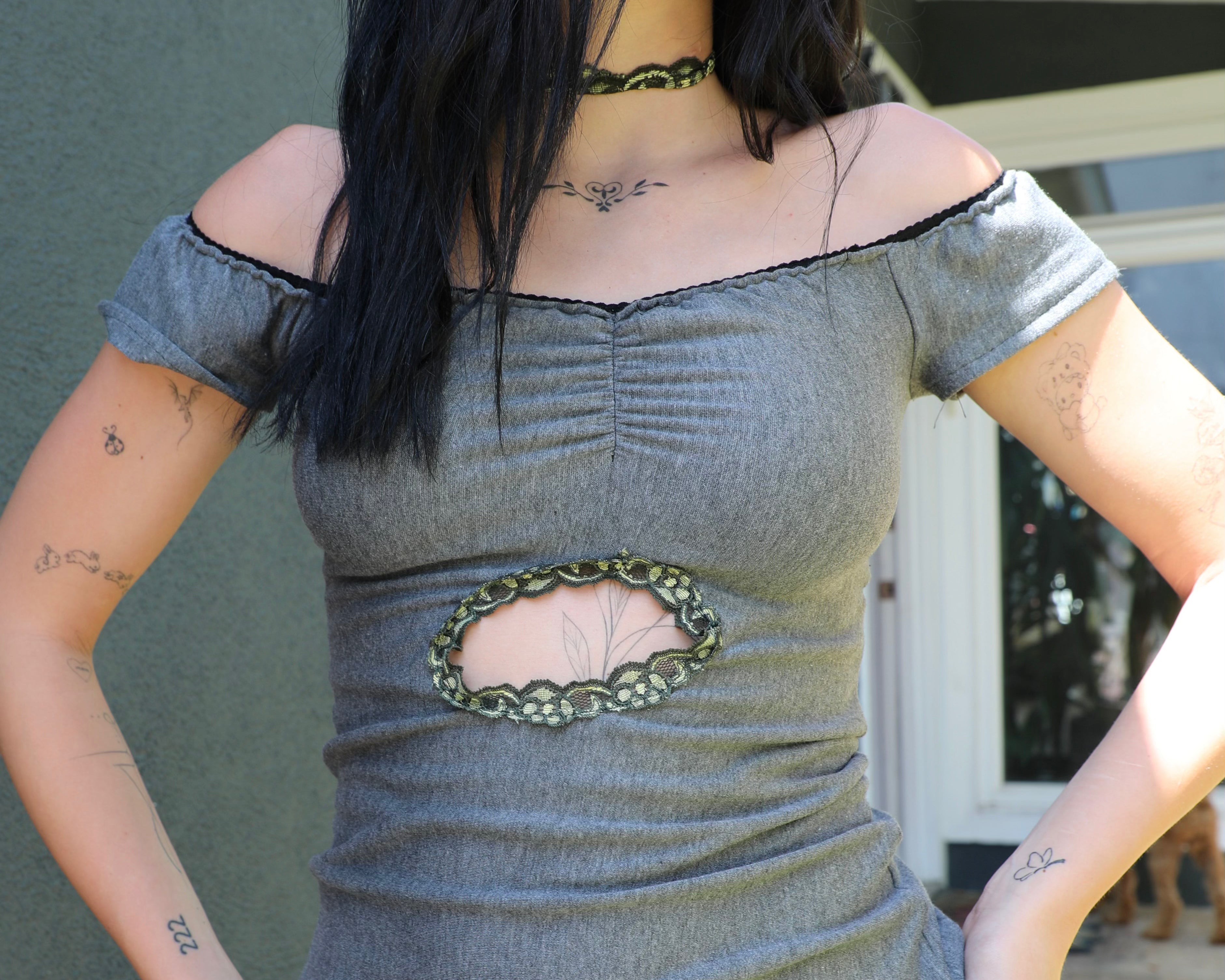 Kai modeling a gray off-the-shoulder top with a central cutout framed by green chain detailing, paired with matching gray shorts featuring a circular patch, photographed outdoors against a building wall.