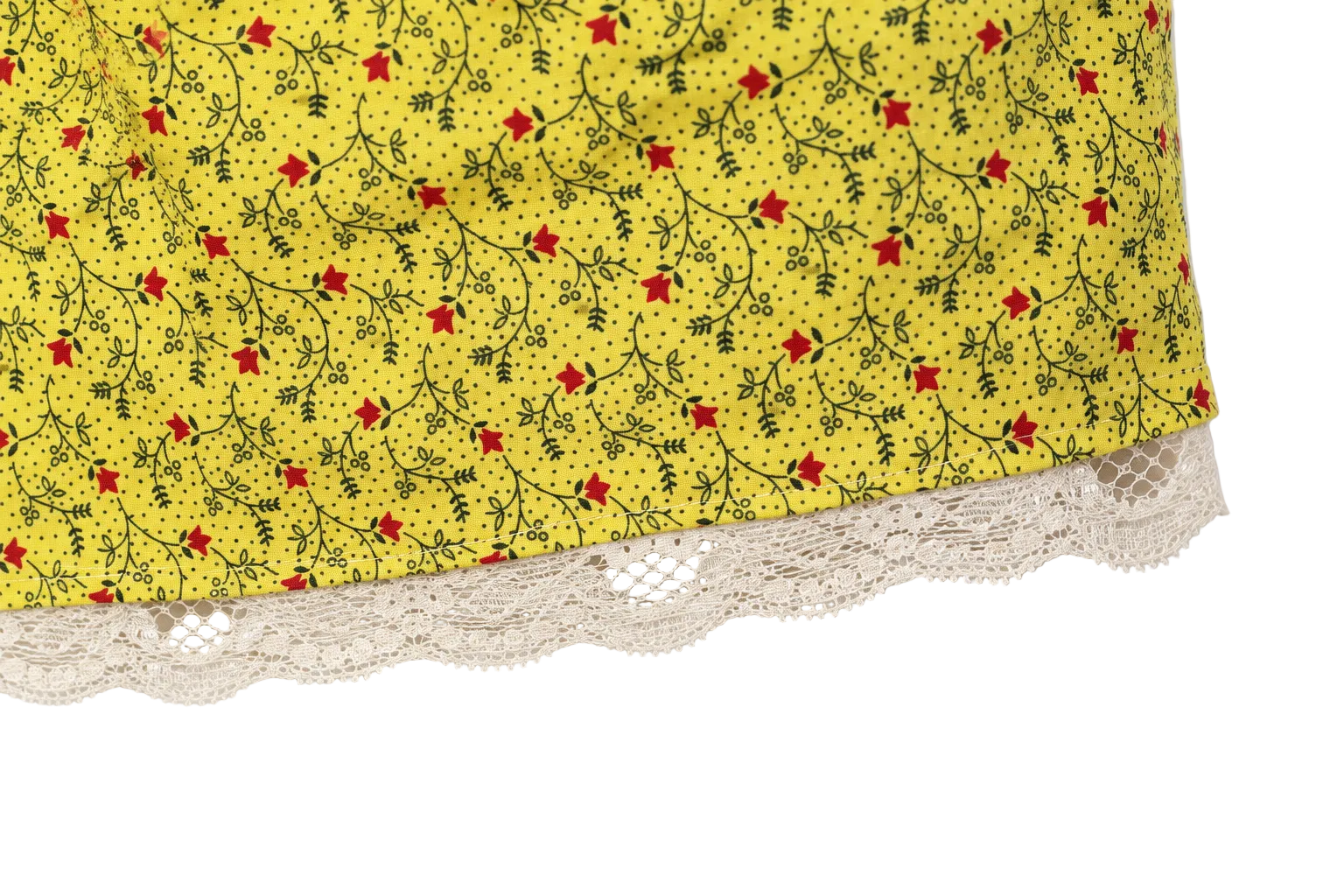 Close-up of a yellow fabric item with a small red and black floral vine print and a cream lace trim along the bottom edge, shown against a white background.