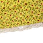Close-up of a yellow fabric item with a small red and black floral vine print and a cream lace trim along the bottom edge, shown against a white background.
