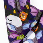 A close-up of a Halloween-themed fabric featuring a repeating pattern of cartoon bats, purple wings, orange trick-or-treat bags, and white moon shapes on a dark background.