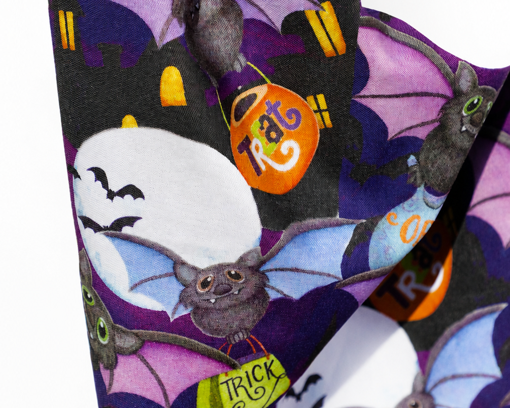 A close-up of a Halloween-themed fabric featuring a repeating pattern of cartoon bats, purple wings, orange trick-or-treat bags, and white moon shapes on a dark background.